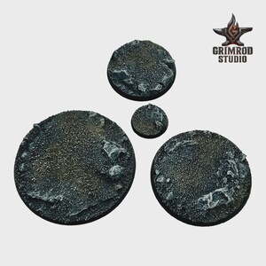 May include: Four textured, circular bases for miniature figures. The bases vary in size, with a rough, rocky surface in shades of gray and brown. The Grimrod Studio logo is in the upper right corner.