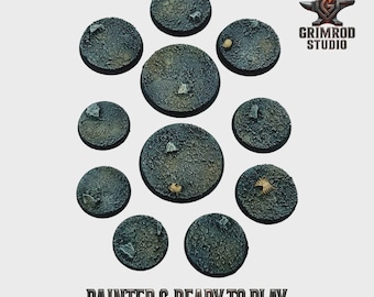 May include: A collection of eleven painted, round miniature bases with a textured, cracked earth appearance. The bases are various sizes and feature small rocks and debris. The text "PAINTED & READY TO PLAY" is at the bottom, with the Grimrod Studio logo in the upper right.