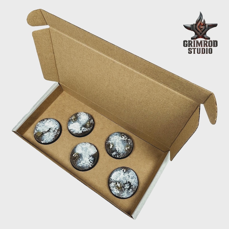 Op de afbeelding: Een bruine kartonnen doos met vijf ronde, getextureerde bases voor miniaturen. De bases hebben een besneeuwd terreinontwerp met witte, grijze en gouden accenten. Het Grimrod Studio-logo is rechtsboven zichtbaar.