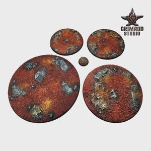 Pu&ograve; includere: Una collezione di basi circolari strutturate di varie dimensioni, progettate per figure in miniatura. Le basi presentano un terreno marrone-rossastro con macchie di pietra grigia e riflessi gialli. Il logo Grimrod Studio &egrave; visibile in alto a destra.