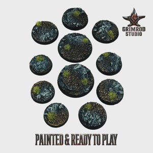 Peut inclure: Dix socles circulaires pour figurines, peints avec un terrain rocheux et des touffes de végétation verte. Les socles sont de tailles variées et prêts à l'emploi. Le texte "PAINTED & READY TO PLAY" est visible.