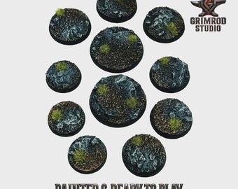 May include: A collection of ten circular, textured bases for miniature figures, painted with a rocky terrain and patches of green vegetation. The bases vary in size and are ready to use. The text "PAINTED & READY TO PLAY" is at the bottom.