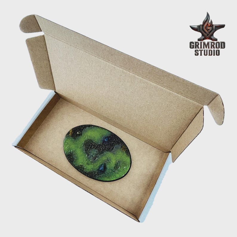 May include: An oval-shaped, green and black decorative item with blue accents, inside a cardboard box. The item has a textured, speckled appearance. The box is open, revealing the product. The Grimrod Studio logo is visible.