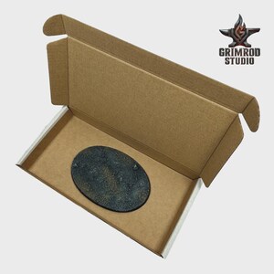 May include: An open cardboard box with a textured, oval-shaped base inside. The base is dark gray with a rough, rocky appearance. The box has a white interior and the logo "GRIMROD STUDIO" is visible in the upper right corner.