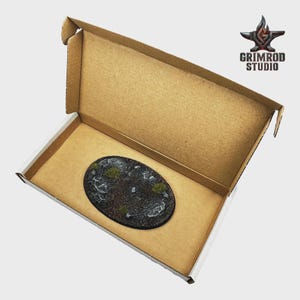 May include: An oval-shaped miniature base with a textured, rocky surface and patches of green vegetation, presented inside an open cardboard box. The base is dark gray and black, with white accents. The Grimrod Studio logo is visible in the upper right corner.