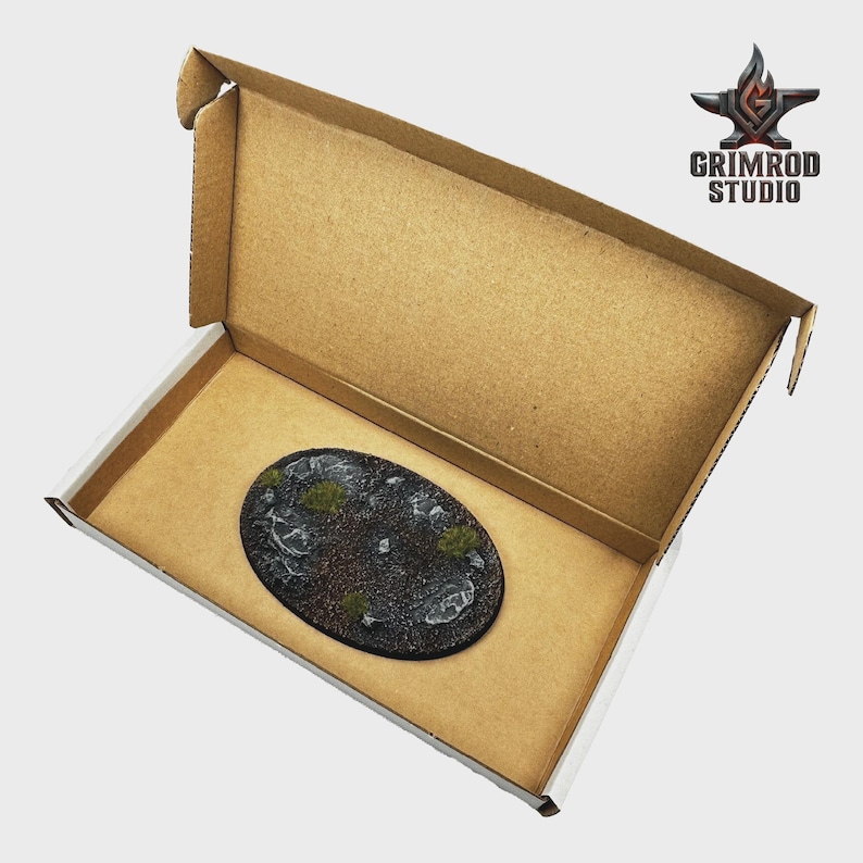 May include: An oval-shaped miniature base with a textured, rocky surface and patches of green vegetation, presented inside an open cardboard box. The base is dark gray and black, with white accents. The Grimrod Studio logo is visible in the upper right corner.