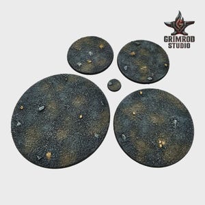 May include: A collection of textured, circular bases in varying sizes, designed for miniature figures. The bases are primarily dark gray with brown and tan accents, and small rock-like details. The Grimrod Studio logo is visible in the upper right corner.