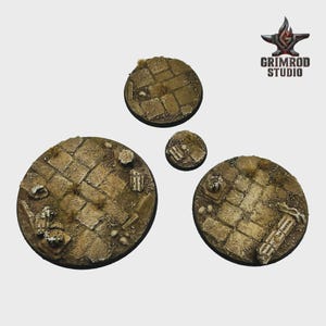 May include: Set of four miniature bases with a cobblestone texture, ideal for tabletop gaming. The bases are circular, with varying diameters, and feature a weathered, aged appearance with small details like grass and debris. The Grimrod Studio logo is in the upper right corner.