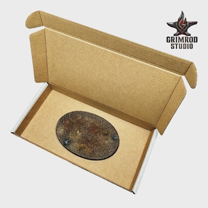 May include: An oval-shaped miniature base with a textured, earthy design, presented inside an open cardboard box. The base features a dark border and is likely for tabletop gaming miniatures. The Grimrod Studio logo is visible.