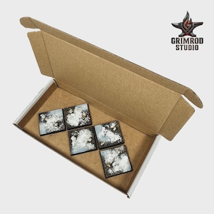 Op de afbeelding: Een open kartonnen doos bevat zes vierkante miniatuur terraintegels. De tegels hebben een donkere basis met witte en gouden accenten, die lijken op een bewolkte hemel. Het Grimrod Studio-logo is rechtsboven zichtbaar.