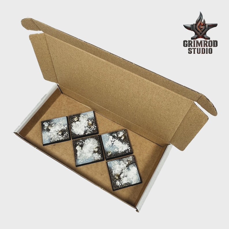 Op de afbeelding: Een open kartonnen doos bevat zes vierkante miniatuur terraintegels. De tegels hebben een donkere basis met witte en gouden accenten, die lijken op een bewolkte hemel. Het Grimrod Studio-logo is rechtsboven zichtbaar.