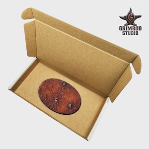 May include: An open cardboard box with a brown oval-shaped object inside. The object has a textured, reddish-brown surface with two small, silver-colored details. The box has the Grimrod Studio logo.