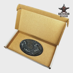 May include: An open cardboard box with a brown interior and white exterior. Inside, an oval, textured, black base is visible. The Grimrod Studio logo is in the upper right corner.
