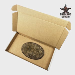 May include: An open cardboard box with a brown interior and a textured, oval-shaped base. The base has a cobblestone design. The Grimrod Studio logo is in the upper right corner.