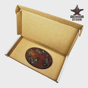 Pu&ograve; includere: Una base in miniatura ovale con una superficie strutturata marrone-rossastra e rocce grigie sparse, presentata all'interno di una scatola di cartone aperta. La scatola ha un interno marrone e un esterno bianco. Il logo Grimrod Studio &egrave; visibile.