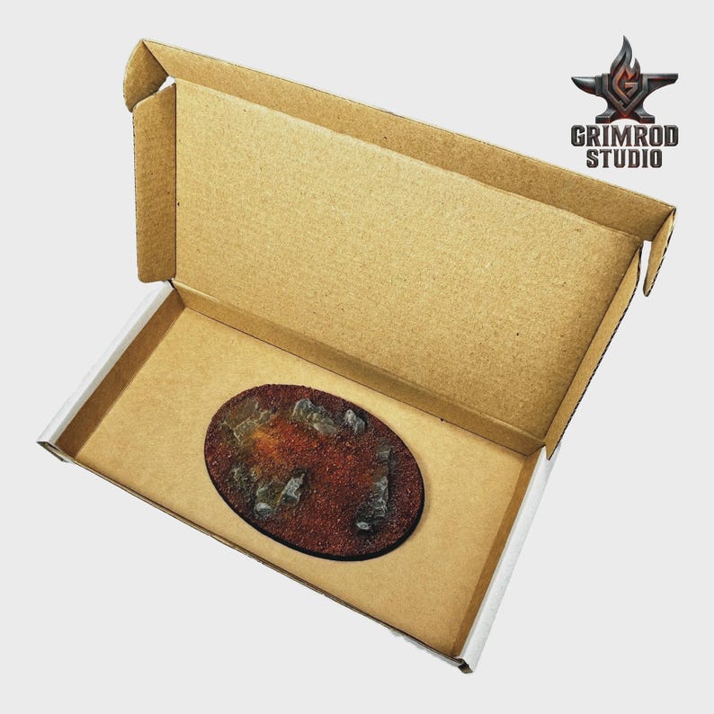 Pu&ograve; includere: Una base in miniatura ovale con una superficie strutturata marrone-rossastra e rocce grigie sparse, presentata all'interno di una scatola di cartone aperta. La scatola ha un interno marrone e un esterno bianco. Il logo Grimrod Studio &egrave; visibile.
