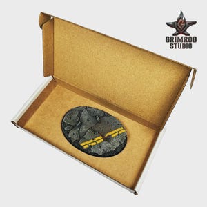 May include: An oval-shaped miniature base with a cracked asphalt design and yellow stripes, presented inside a cardboard box. The Grimrod Studio logo is visible in the upper right corner.