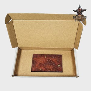 May include: An open cardboard box with a rectangular, reddish-brown item inside. The item has a textured surface with white speckles. The box has the text "GRIMROD STUDIO" and a logo on the top right corner.