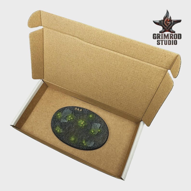 May include: An oval-shaped miniature base with a textured, dark green surface, featuring small, light green and gray elements, is presented inside a cardboard box. The box has a brown exterior and white interior. The Grimrod Studio logo is visible.