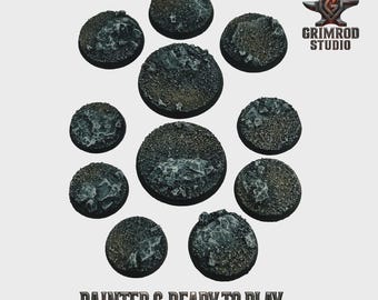 May include: A collection of eleven round, textured bases, painted in shades of gray and brown, arranged in a circular pattern. The bases have a rocky appearance with lighter gray accents. The text "PAINTED & READY TO PLAY" is at the bottom, with the Grimrod Studio logo in the upper right.