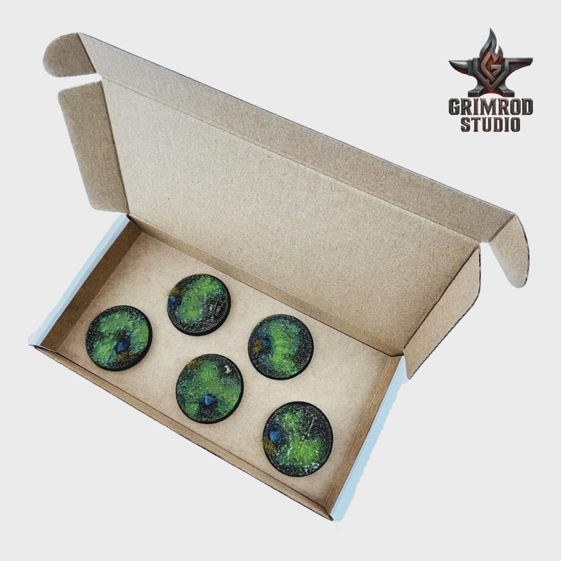 May include: An open cardboard box containing five circular, textured bases with green and black detailing. The bases are arranged inside the box. The Grimrod Studio logo is visible in the upper right corner.