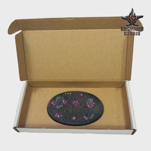 May include: An oval, black-painted base with purple floral accents, presented inside an open cardboard box. The base features a textured surface and is decorated with small, raised purple flowers and green leaves. The box has the text "GRIMROD STUDIO" and a logo.
