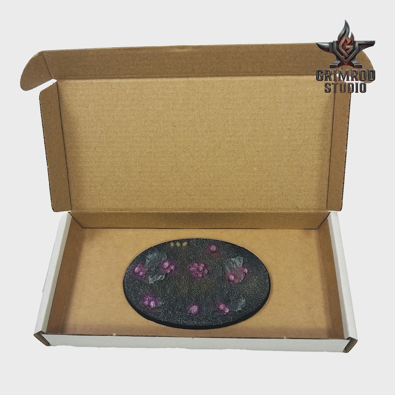May include: An oval, black-painted base with purple floral accents, presented inside an open cardboard box. The base features a textured surface and is decorated with small, raised purple flowers and green leaves. The box has the text "GRIMROD STUDIO" and a logo.