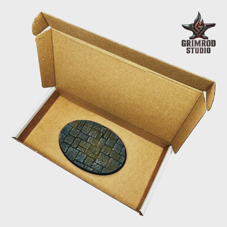 May include: An oval-shaped miniature base with a cobblestone design sits inside an open cardboard box. The base features a detailed pattern of gray and green stones. The box is brown with white edges and the Grimrod Studio logo is visible.