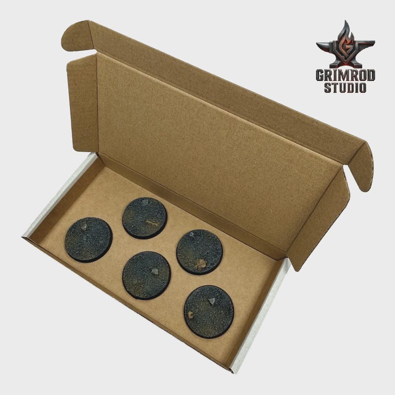 May include: A cardboard box with a white interior holds five round, textured bases. The bases are dark with a mottled appearance, suggesting a terrain or weathered effect. The Grimrod Studio logo is visible.