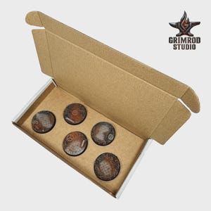 Pu&ograve; includere: Scatola di cartone aperta contenente cinque basi circolari strutturate. Le basi presentano un design industriale arrugginito con accenti metallici. Il logo Grimrod Studio &egrave; visibile in alto a destra.