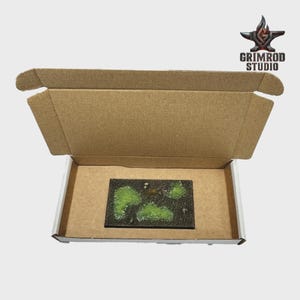 May include: A rectangular terrain base with green and white accents, presented inside an open cardboard box. The base features a dark, textured surface with irregular green shapes. The box has the text "GRIMROD STUDIO" and a logo.