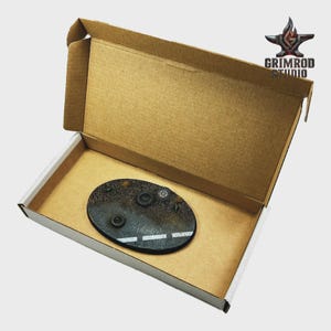May include: An oval, textured, black base with a white road marking and several small, dark, circular objects. The base is inside a brown cardboard box with the Grimrod Studio logo.