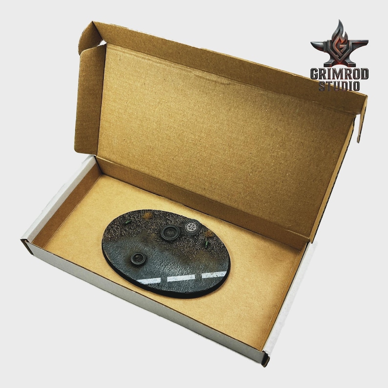 May include: An oval, textured, black base with a white road marking and several small, dark, circular objects. The base is inside a brown cardboard box with the Grimrod Studio logo.