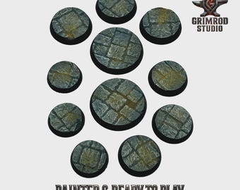 May include: A collection of eleven circular, stone-textured bases, painted in shades of gray and brown, arranged on a light background. The bases vary in size and feature a cobblestone pattern. The text "PAINTED & READY TO PLAY" is at the bottom.
