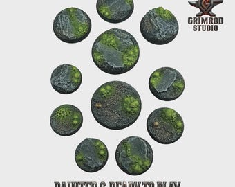 May include: A collection of ten circular miniature bases, painted with a textured, rocky terrain and patches of green moss. The bases are arranged in a circular pattern. The text "PAINTED & READY TO PLAY" is at the bottom.