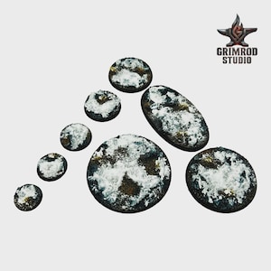 May include: A collection of miniature bases in various shapes, including round and oval, with a textured, dark brown surface and patches of white. The Grimrod Studio logo is in the upper right corner.