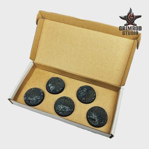 May include: A cardboard box containing five black, textured, circular bases. The bases have a rough, rocky appearance. The box has the Grimrod Studio logo on the upper right corner.