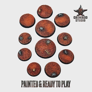 May include: A collection of eleven round, textured bases, painted in shades of brown and orange, with small rock details. The bases are arranged in a circular pattern. The text "PAINTED & READY TO PLAY" is at the bottom.