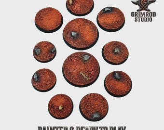 May include: A collection of eleven round, textured bases, painted in shades of brown and orange, with small rock details. The bases are arranged in a circular pattern. The text "PAINTED & READY TO PLAY" is at the bottom.