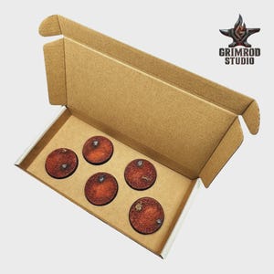 May include: A cardboard box containing five round, textured bases with a reddish-brown color and small metallic accents. The bases are arranged inside a white-lined compartment. The Grimrod Studio logo is visible in the upper right corner.