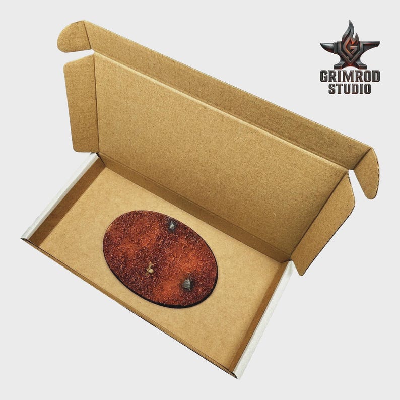 May include: An open cardboard box with a brown oval-shaped item inside. The item has a textured, reddish-brown surface with two small, silver-colored details. The box has the Grimrod Studio logo.
