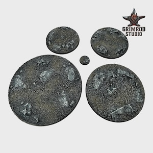 May include: A collection of textured, circular bases in varying sizes, designed for miniature figures. The bases have a dark, speckled surface with embedded, irregular, light-colored rock formations. The Grimrod Studio logo is visible in the upper right corner.