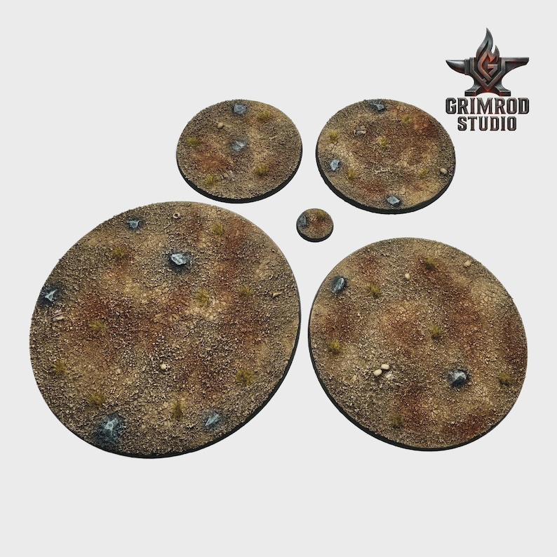 May include: A collection of textured, circular bases for miniature figures. The bases are various sizes, with a brown, sandy texture, small rocks, and sparse green vegetation. The Grimrod Studio logo is in the upper right corner.