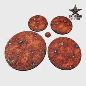 May include: A collection of textured, circular bases in varying sizes, featuring a reddish-brown color scheme with embedded metallic and stone-like details. The bases are designed for miniature figures, with the Grimrod Studio logo visible in the upper right corner.