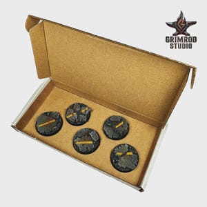May include: A cardboard box containing five circular, textured bases. The bases are dark gray with yellow stripes. The Grimrod Studio logo is visible in the upper right corner.