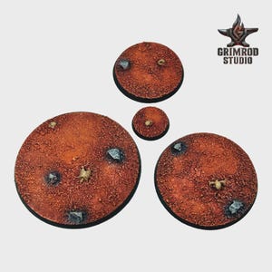 May include: Four circular miniature bases with a textured, reddish-brown surface. Each base features small rocks and a miniature skeleton. The bases vary in size, with the largest measuring approximately 2 inches in diameter. The Grimrod Studio logo is in the upper right corner.