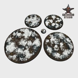 May include: A collection of miniature bases with a snow-covered terrain effect. The bases are various sizes, ranging from small circles to larger ovals. The bases are brown with white and gray accents, and the Grimrod Studio logo is in the upper right corner.