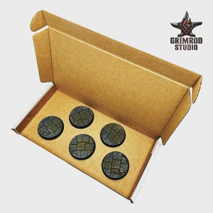 May include: A cardboard box containing five circular, textured bases with a cobblestone pattern. The bases are dark gray and green. The Grimrod Studio logo is visible in the upper right corner.