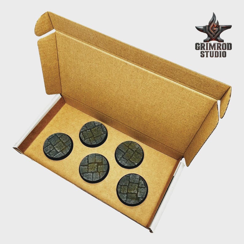 May include: A cardboard box containing five circular, textured bases with a cobblestone pattern. The bases are dark gray and green. The Grimrod Studio logo is visible in the upper right corner.