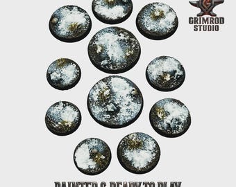 May include: A collection of eleven circular, textured bases for miniature figures. The bases are painted with a snow and grass effect, with white and green tones. The text "PAINTED & READY TO PLAY" is displayed at the bottom.